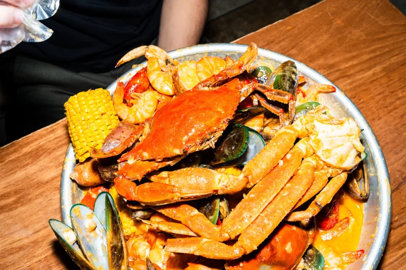 A metal bowl piled with a whole blue crab, snow crab legs, shrimp, green mussels, corn on the cob, sausage, and crawfish in Big Crab sauce